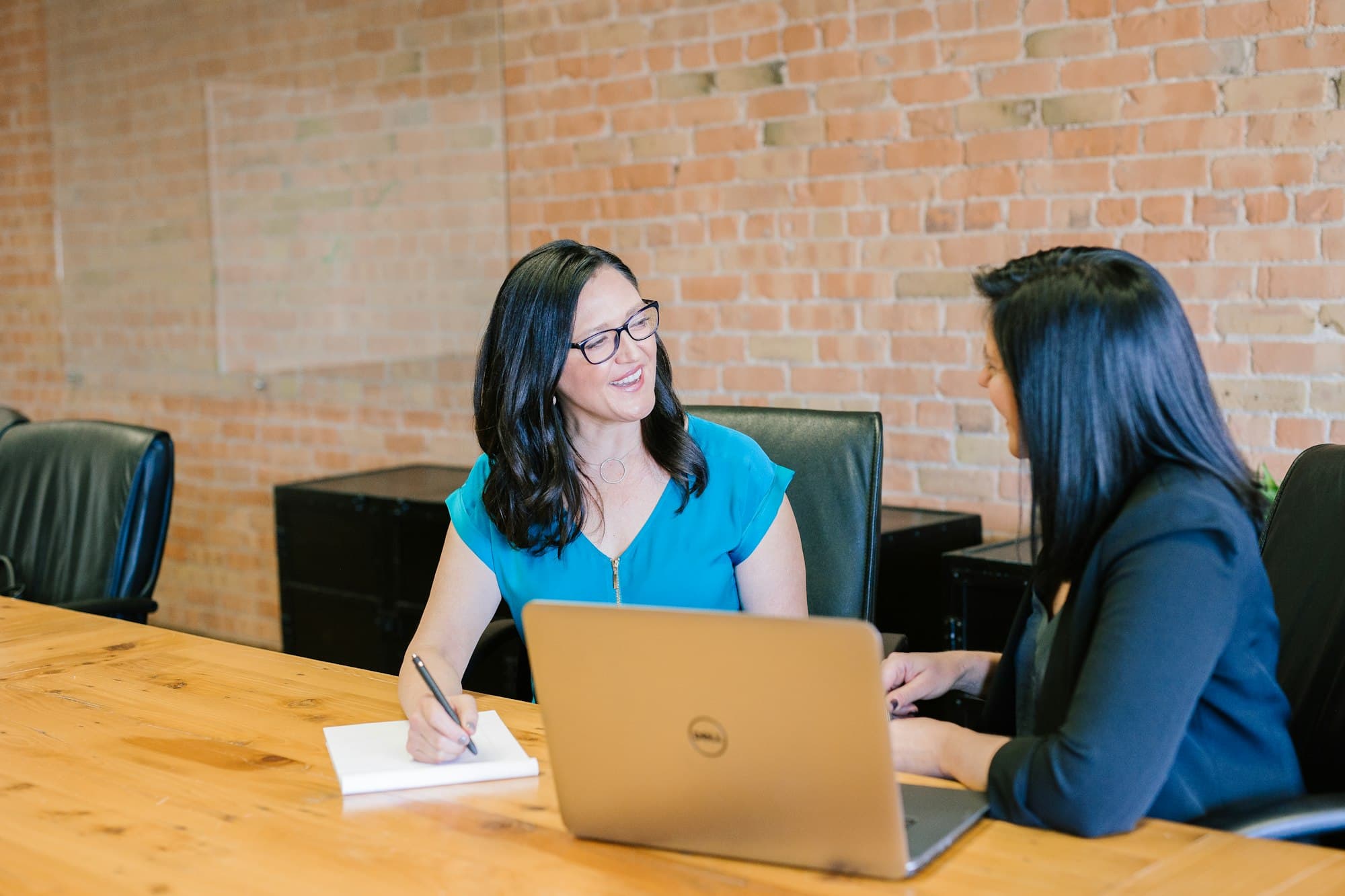 Two women talking across a table with a laptop and notepad, one explaining something to the other
